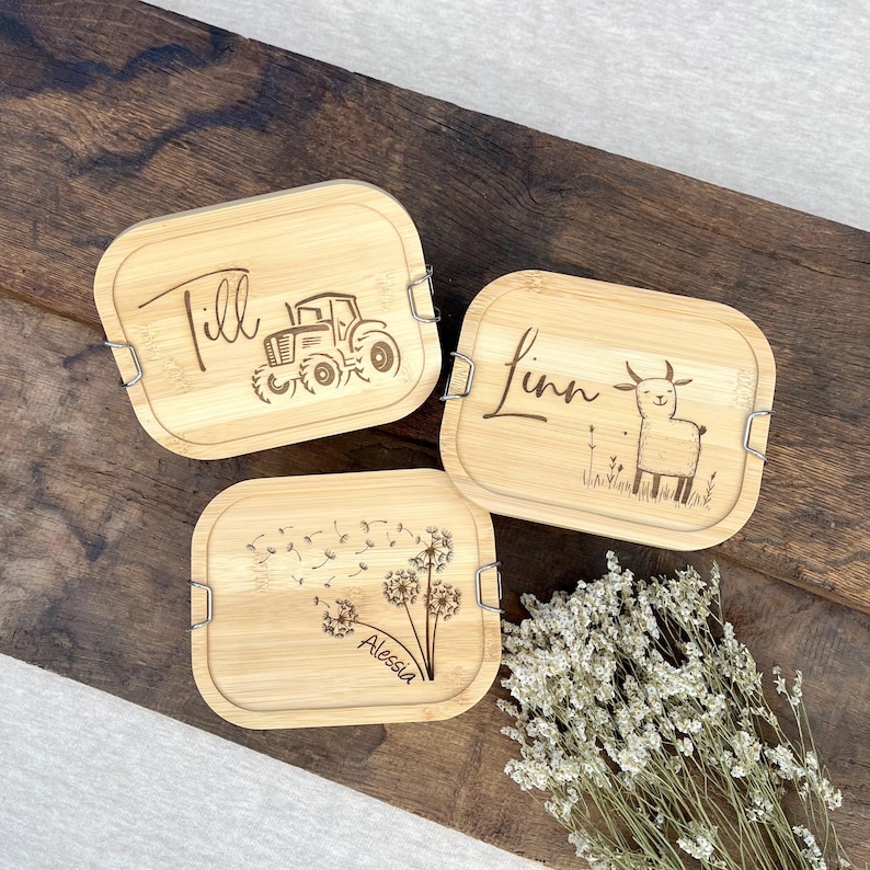 Drei rechteckige Bambus-Lunchboxen mit silbernen Verschlüssen. Jeder Deckel ist mit einem Namen und einem Design graviert: ein Traktor, eine Ziege und ein Löwenzahn. Die Namen sind Till, Linn und Alessia.