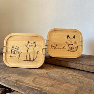 May include: Two rectangular bamboo lunch boxes with rounded corners and metal side clips. One lid features a laser-engraved cat and the name Lilly, while the other lid displays a laser-engraved fox and the name Paul in black ink.