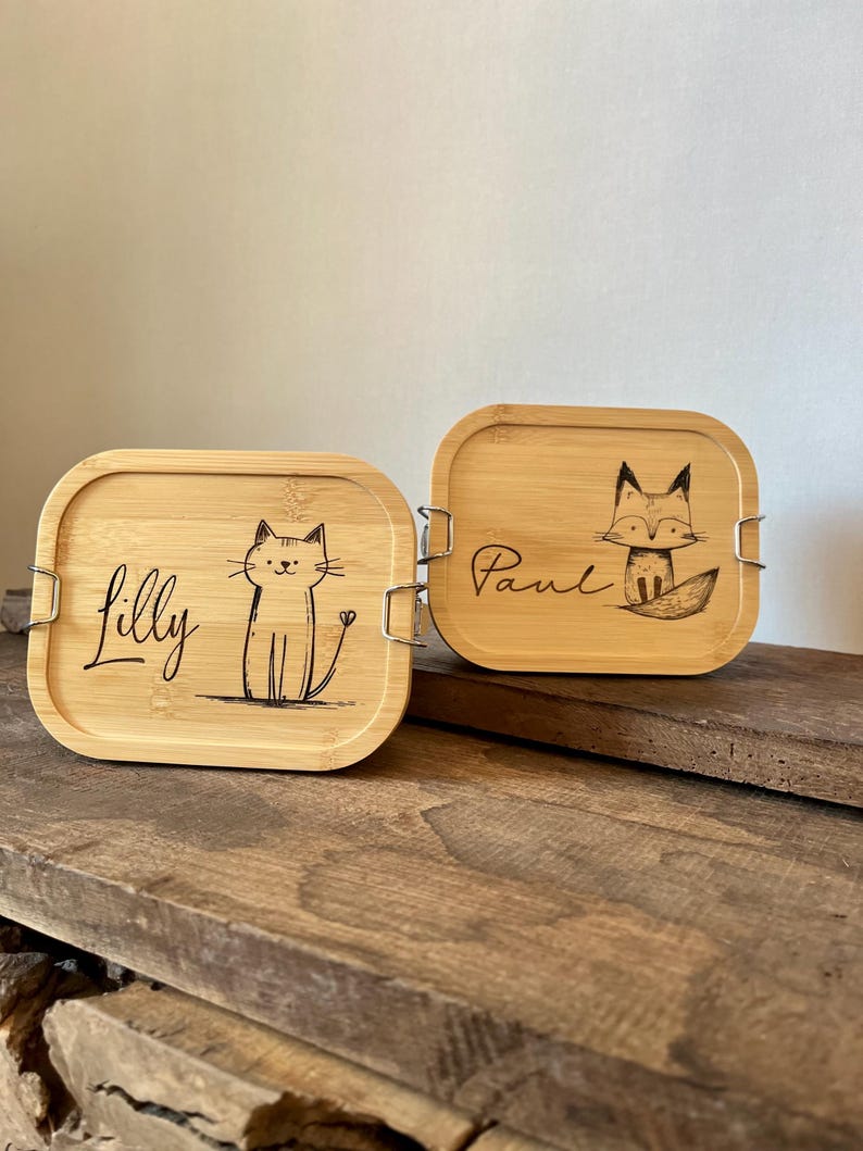 May include: Two rectangular bamboo lunch boxes with rounded corners and metal side clips. One lid features a laser-engraved cat and the name Lilly, while the other lid displays a laser-engraved fox and the name Paul in black ink.