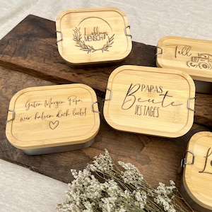 Eine Sammlung von Bambus-Lunchboxen mit silbernen Verschlüssen. Die Deckel sind mit Text und Designs graviert, darunter die Sätze "Lieblings Mensch", "Guten Morgen Papa, wir haben dich lieb!" mit einem Herz und "Papas Beute des Tages."