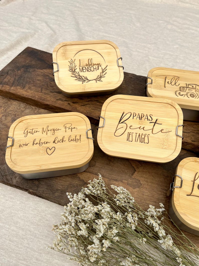 Eine Sammlung von Bambus-Lunchboxen mit silbernen Verschlüssen. Die Deckel sind mit Text und Designs graviert, darunter die Sätze "Lieblings Mensch", "Guten Morgen Papa, wir haben dich lieb!" mit einem Herz und "Papas Beute des Tages."