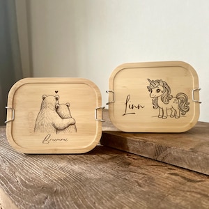 May include: Two rectangular bamboo lunch boxes with rounded corners and metal side latches. One box features a laser-engraved illustration of two bears hugging with the name Brummi, and the other shows a unicorn with the name Linn.