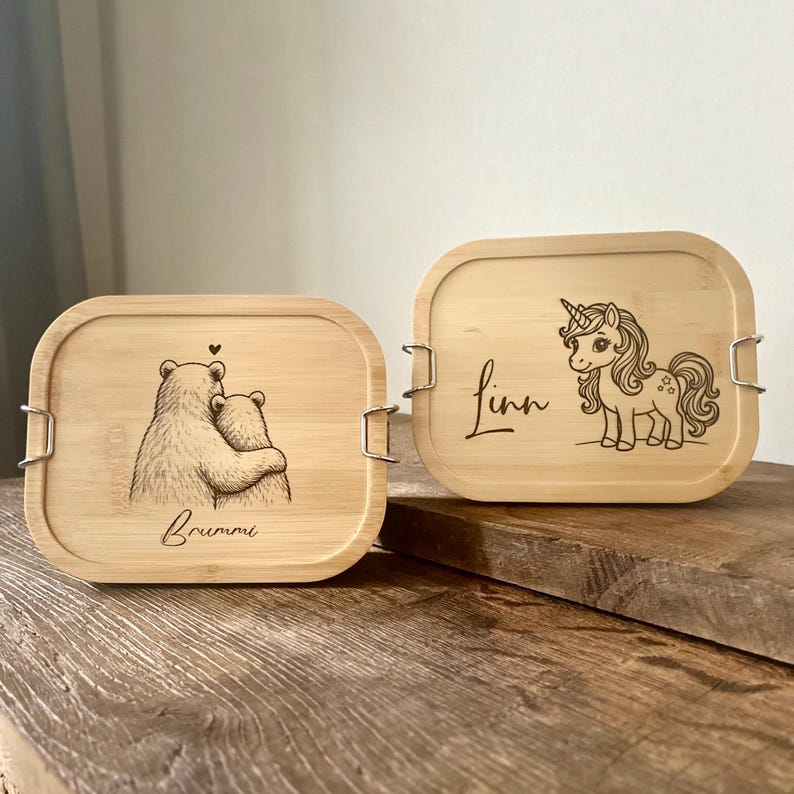 May include: Two rectangular bamboo lunch boxes with rounded corners and metal side latches. One box features a laser-engraved illustration of two bears hugging with the name Brummi, and the other shows a unicorn with the name Linn.