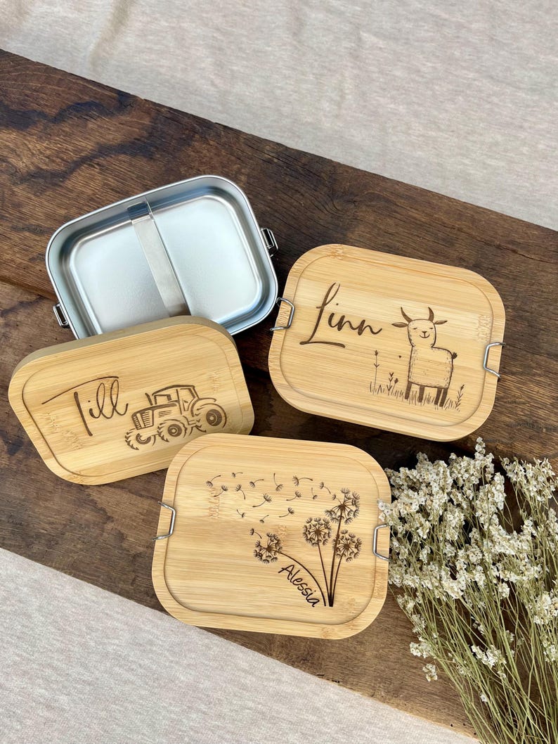 Eine Edelstahl-Lunchbox mit Bambusdeckel. Die Deckel sind mit Namen und Illustrationen graviert: ein Traktor, ein Lama und ein Löwenzahn. Die Lunchbox ist geöffnet und gibt den Innenraum frei. Der Hintergrund ist eine dunkle Holzoberfläche.