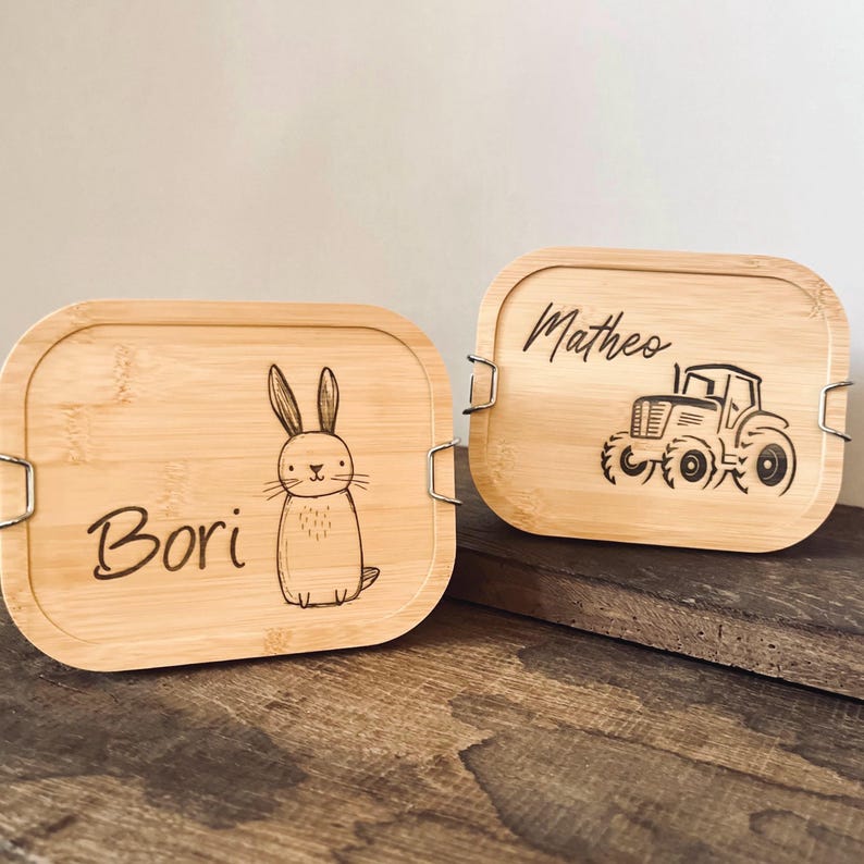 May include: Two rectangular bamboo lunch boxes with rounded corners and metal side clips. One lid features a laser-engraved rabbit and the name Bori, while the other lid displays a laser-engraved tractor and the name Matheo in script lettering.