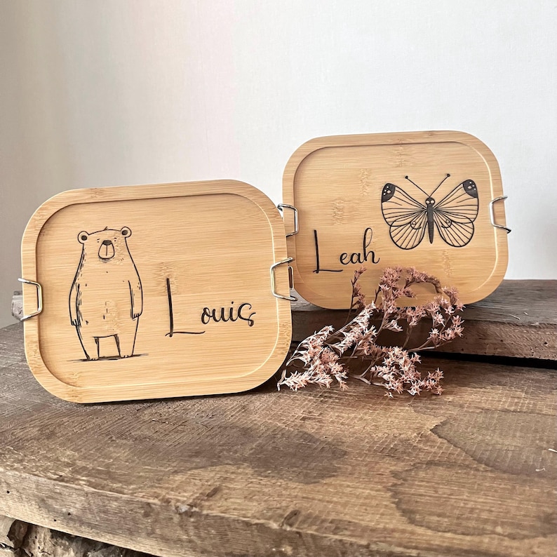 May include: Two rectangular bamboo lids with rounded corners and metal side clips. One lid features a laser-engraved bear and the name Louis. The other lid displays a laser-engraved butterfly and the name Leah, accompanied by dried flowers.