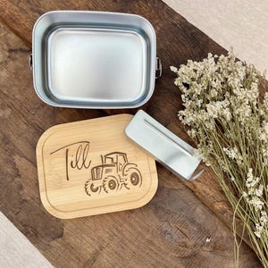 Eine silberne Metall-Lunchbox mit einem Bambusdeckel, auf dem der Name "Till" und ein Traktormotiv eingraviert sind. Ein silbernes Metallutensil liegt neben der Lunchbox. Getrocknete Blumen liegen auf einer Holzoberfläche.