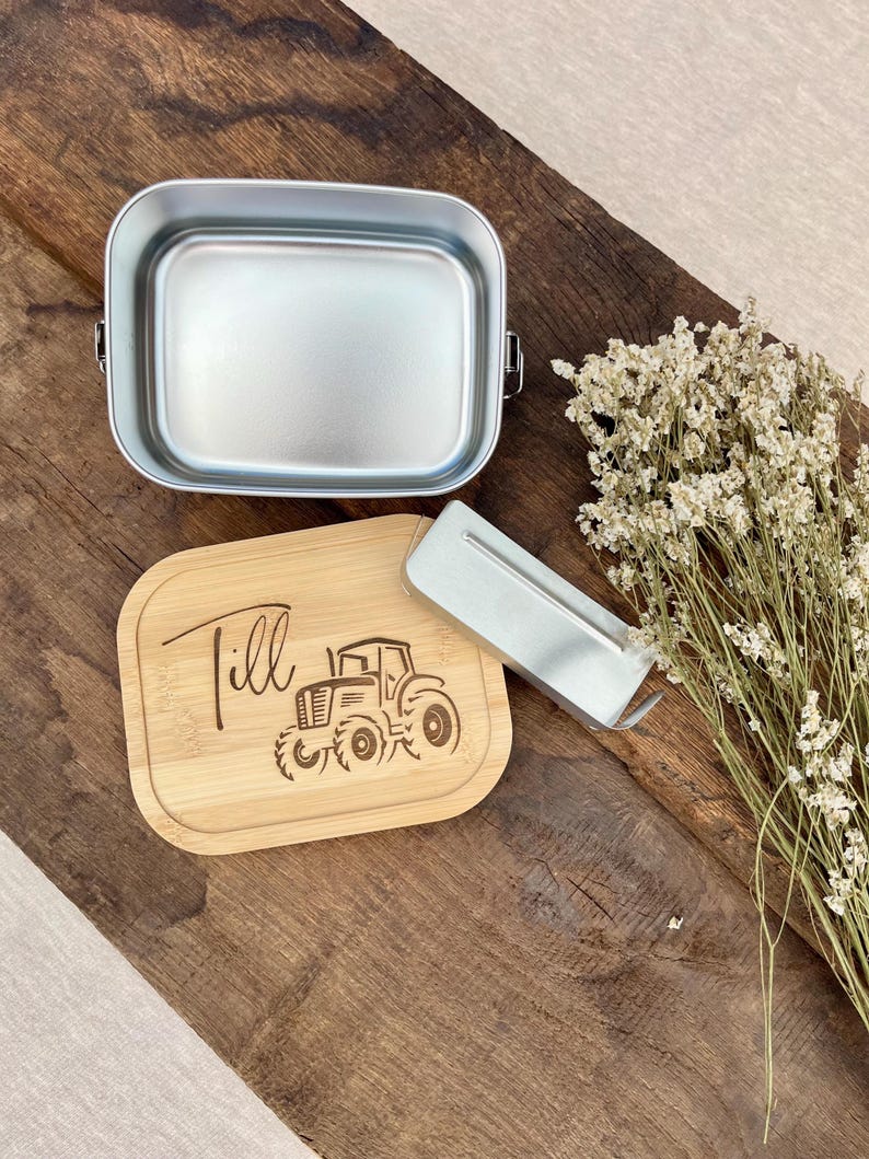 Eine silberne Metall-Lunchbox mit einem Bambusdeckel, auf dem der Name "Till" und ein Traktormotiv eingraviert sind. Ein silbernes Metallutensil liegt neben der Lunchbox. Getrocknete Blumen liegen auf einer Holzoberfläche.