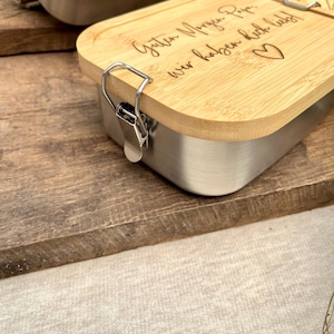 Edelstahl-Lunchbox mit Bambusdeckel. Der Deckel ist mit den Worten "Guten Morgen Papa, wir haben dich lieb!" und einem Herzsymbol graviert. Die Box hat einen silbernen Verschluss und steht auf einer Holzoberfläche.