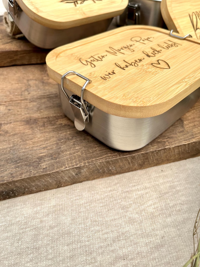 Edelstahl-Lunchbox mit Bambusdeckel. Der Deckel ist mit den Worten "Guten Morgen Papa, wir haben dich lieb!" und einem Herzsymbol graviert. Die Box hat einen silbernen Verschluss und steht auf einer Holzoberfläche.