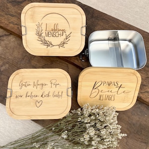 Ein Set aus vier Bambus-Lunchboxen mit Edelstahl-Innenräumen. Drei Deckel sind mit deutschem Text und dekorativen Elementen graviert. Ein Deckel trägt die Aufschrift "Lieblingsmensch" in einem Kranz. Die anderen beiden lauten "Guten Morgen Papa, wir haben dich lieb!" und "PAPAS Beute des Tages".