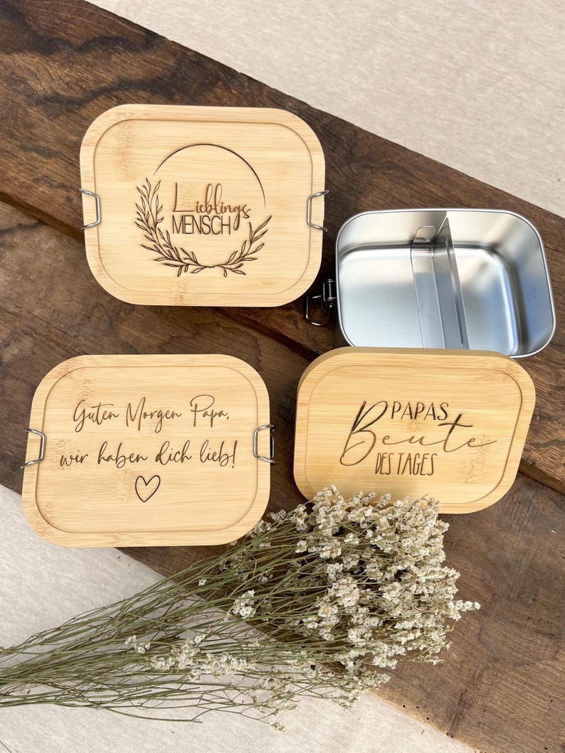 Ein Set aus vier Bambus-Lunchboxen mit Edelstahl-Innenräumen. Drei Deckel sind mit deutschem Text und dekorativen Elementen graviert. Ein Deckel trägt die Aufschrift "Lieblingsmensch" in einem Kranz. Die anderen beiden lauten "Guten Morgen Papa, wir haben dich lieb!" und "PAPAS Beute des Tages".