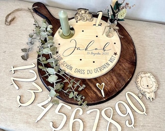 May include: Wooden board with a round, light wood center engraved with the name "Jakob" and the date "19 Dezember 2023" and the text "Wie schön, dass du geboren bist". The board is surrounded by a dark wood frame. There are wooden numbers 1 through 9, a small wooden tractor, a small wooden lion, and a small glass jar with a sprig of greenery.