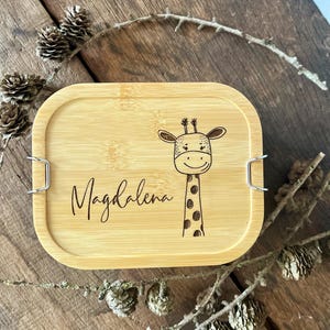 May include: A rectangular bamboo lunch box lid featuring a laser-engraved illustration of a giraffe and the name Magdalena in cursive script. The lid has rounded corners and two silver-colored metal side clips for securing the container.