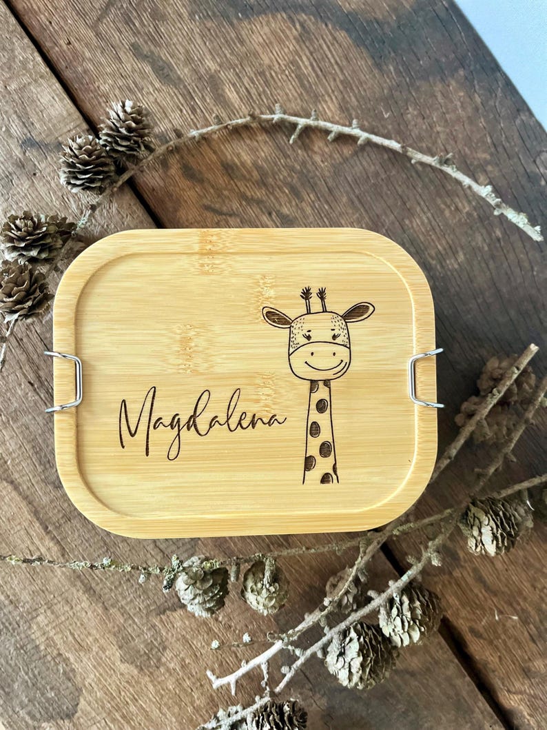 May include: A rectangular bamboo lunch box lid featuring a laser-engraved illustration of a giraffe and the name Magdalena in cursive script. The lid has rounded corners and two silver-colored metal side clips for securing the container.