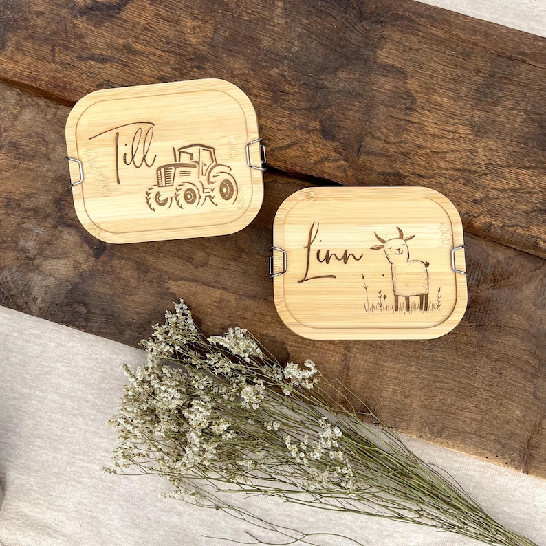 Zwei Bambus-Lunchboxen mit eingravierten Designs. Eine zeigt den Namen "Till" und einen Traktor, die andere den Namen "Linn" und eine Ziegenillustration. Beide haben Metallverschlüsse und liegen auf einer Holzoberfläche.