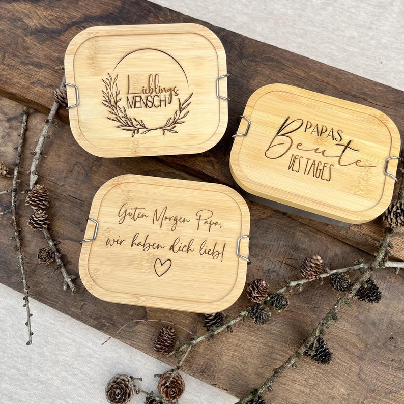 Drei rechteckige Bambus-Lunchboxen mit graviertem Text und silbernen Verschlüssen. Die Deckel zeigen Sätze wie "Lieblingsmensch", "Papas Beute des Tages" und "Guten Morgen Papa, wir haben dich lieb!" mit einem Herz. Auf einer Holzoberfläche.