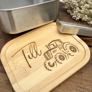 Ein helles Holzschneidebrett mit abgerundeten Kanten, graviert mit dem Namen "Till" und einem Traktormotiv. Ein Edelstahlbehälter und eine Zange sind ebenfalls vorhanden.