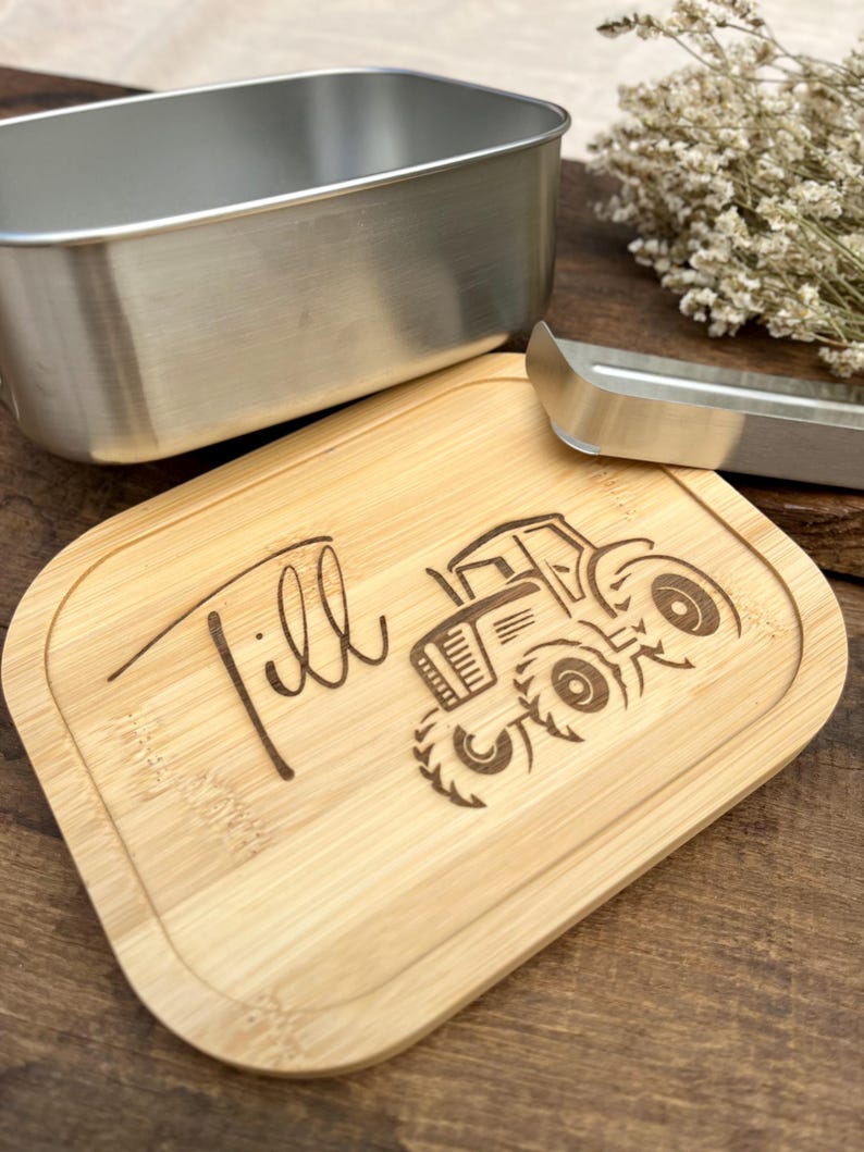 Ein helles Holzschneidebrett mit abgerundeten Kanten, graviert mit dem Namen "Till" und einem Traktormotiv. Ein Edelstahlbehälter und eine Zange sind ebenfalls vorhanden.