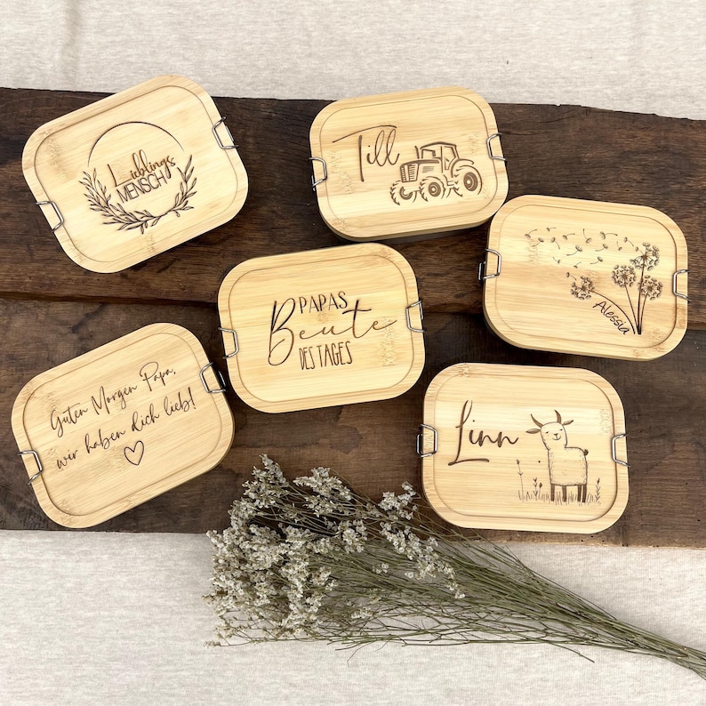 Sechs rechteckige Bambus-Lunchboxen mit eingravierten Designs und Text. Die Designs umfassen einen Traktor, ein Lama, einen Kranz und Pusteblumen. Der Text enthält Namen und Sätze wie "Guten Morgen Papa, wir lieben dich!" Das sind Geschenke