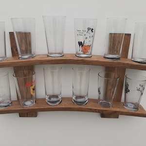 May include: A wooden rack displaying twelve clear pint glasses. The glasses are arranged on two shelves, with some featuring decorative designs. The rack is made of light brown wood and is mounted on a white wall.