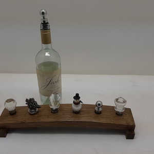 May include: A wooden wine bottle stopper rack with six bottle stoppers. The stoppers are made of glass and metal and feature various designs, including a snowman, a grapevine, and a crystal.