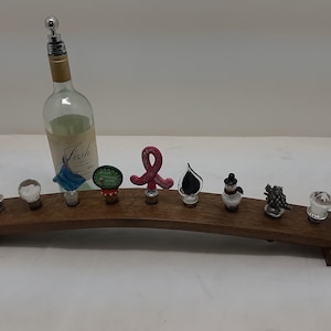 May include: A wooden wine rack with eight decorative bottle stoppers and a wine bottle with a silver spout. The stoppers have various designs, including a snowman, grapes, and a ribbon. The wine bottle has a label that says "Josh".