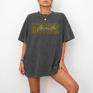 Puede incluir: Una camiseta gris carbón de corte oversize con la palabra MOTHER en una fuente de bloque de color amarillo mostaza. Superpuesto en el centro, el nombre Amanda está escrito en una fina letra cursiva blanca. Tiene mangas cortas y cuello redondo.