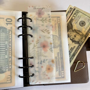 May include: A brown faux leather binder with black rings, holding cash and floral-patterned dividers. The word "Savings" is printed on a divider. The binder also contains a gold-colored paper clip.