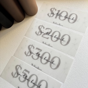 May include: Four translucent gift cards with the text "$100 in the bank", "$200 in the bank", "$300 in the bank", and "$500 in the bank" in a script font. The gift cards have a marble-like pattern. The brand name is at the bottom.