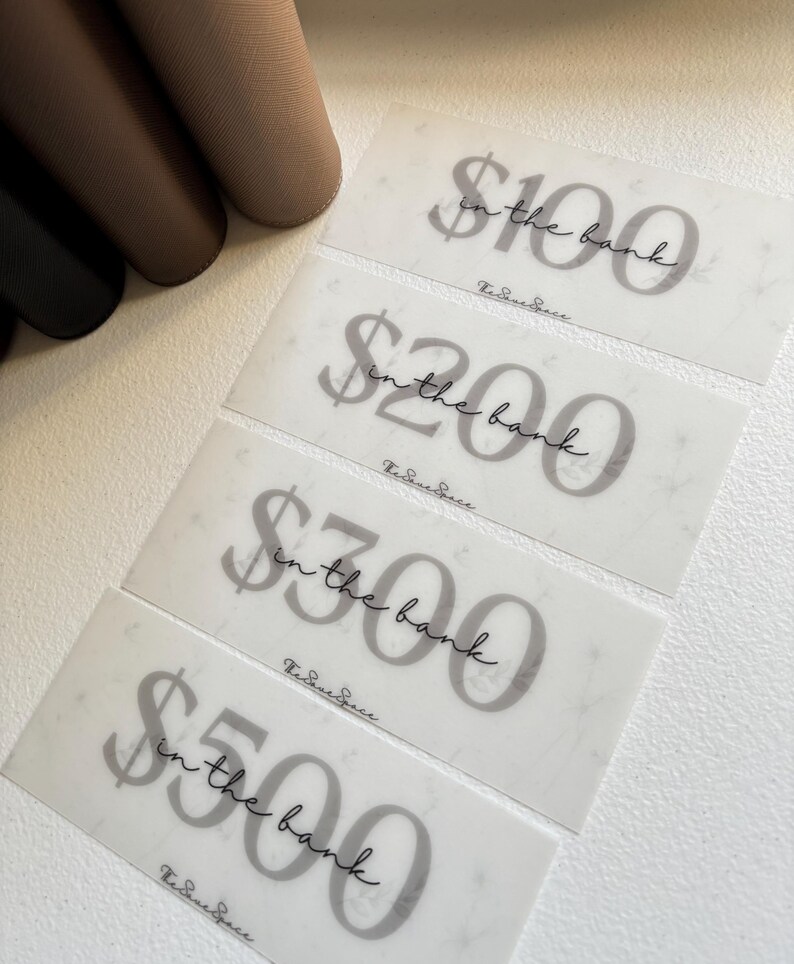 May include: Four translucent gift cards with the text "$100 in the bank", "$200 in the bank", "$300 in the bank", and "$500 in the bank" in a script font. The gift cards have a marble-like pattern. The brand name is at the bottom.