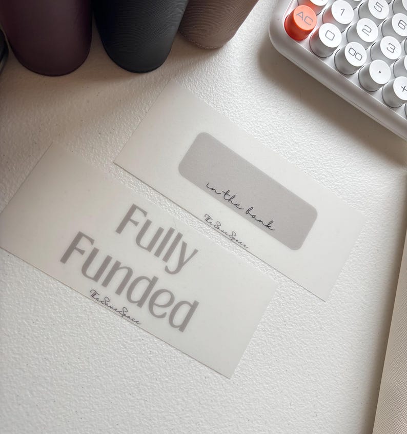 May include: Two white envelopes with gray text. One reads "Fully Funded" in large letters, and the other says "in the bank." Both have a signature-style logo. A calculator and other office supplies are in the background.