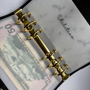 May include: Black binder with gold hardware, holding a fifty-dollar bill and a clear page labeled "Utilities." The clear page has a floral design. The binder is open, revealing the contents.
