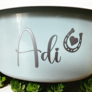 Könnte beinhalten: Eine hellblaue Plastikschüssel mit einem silbernen Vinyl-Aufkleber, auf dem "Adi" mit einem Hufeisen und einem Herz steht.