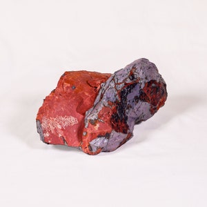 May include: A rough, irregularly shaped rock with a red and purple color pattern. The rock has a smooth, polished surface on one side and a rough, textured surface on the other side.