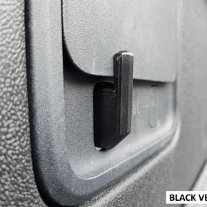 May include: A close-up of a black plastic latch on a gray car door panel. The latch is rectangular and has a small handle. The panel is textured and has a black plastic trim around the edge.