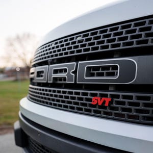 May include: Close-up of a white Ford truck grille. The black grille features the Ford logo in silver and the red SVT emblem. The truck's headlight is visible on the right side of the image.