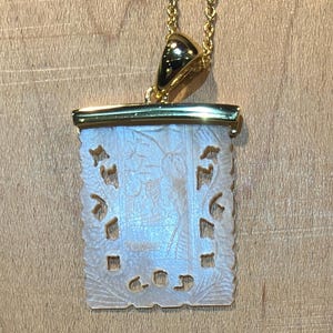 May include: A gold-toned necklace with a rectangular pendant. The pendant features a carved, off-white design with floral and figure motifs. The necklace hangs on a delicate gold chain, creating an elegant accessory.