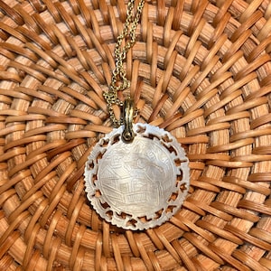 May include: A gold-toned necklace with a circular pendant. The pendant is white with an etched design of a scene with figures and buildings. The pendant has a scalloped edge. The necklace is resting on a woven, brown surface.