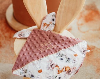 May include: A square baby security blanket featuring a dusty rose dimpled minky fabric on one side and a white cotton fabric with a floral, deer, and hot air balloon print on the other. The text Mon Doudou is embroidered in the center.