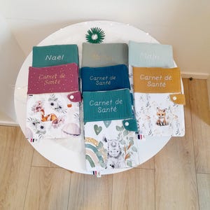 Peut inclure: Six carnets de santé personnalisés pour bébé avec des couvertures de couleurs différentes et des noms brodés. Les couvertures sont en tissu doux et texturé. Chaque carnet a une poche sur le devant pour un stylo ou d'autres petits objets. Les carnets sont décorés d'illustrations d'animaux variés et du texte "Carnet de Santé".