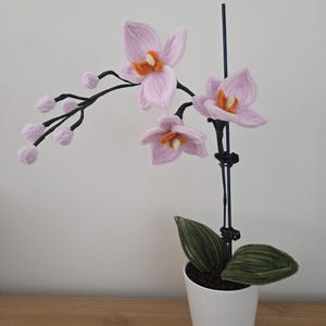 May include: A handcrafted, light pink orchid plant with orange centers and green leaves in a white pot. The artificial flowers are on a black stem. The plant is displayed on a wooden surface against a white wall.