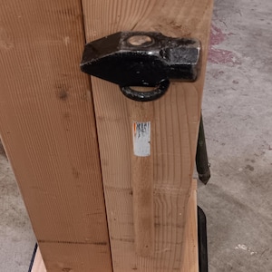 Puede incluir: Un mazo negro con mango de madera está fijado a una estructura de madera. La cabeza del martillo es de metal y tiene un lazo. La madera tiene un veteado natural y está unida con tornillos.