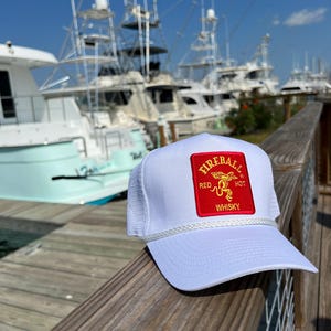 May include: White mesh trucker hat with a red patch that says "Fireball Red Hot Whisky" in gold lettering. The hat is sitting on a wooden railing with a background of boats in a marina.