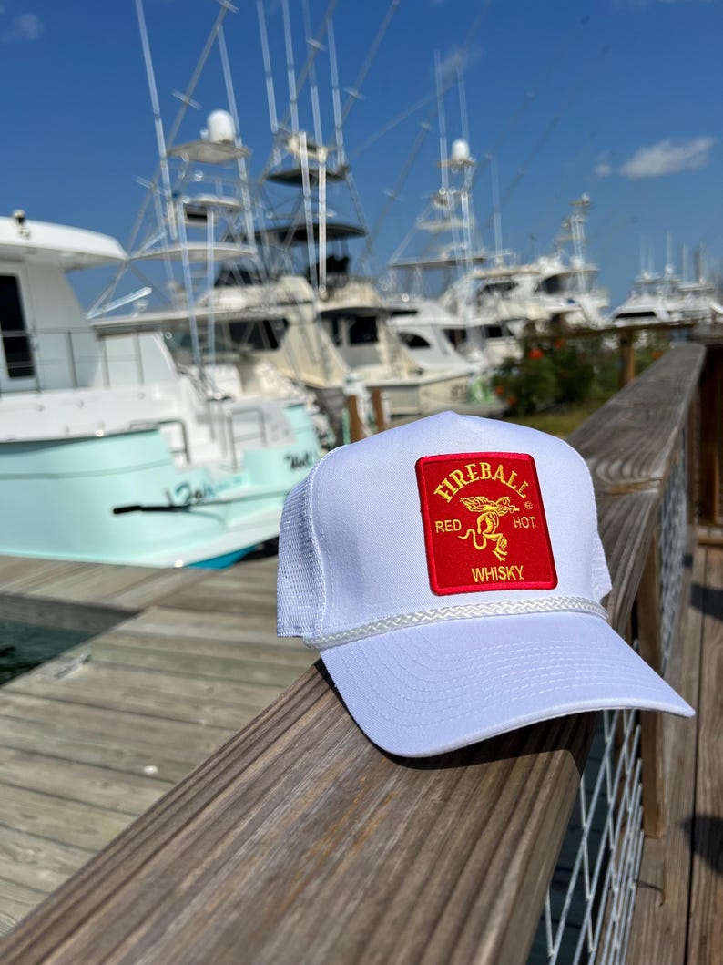 May include: White mesh trucker hat with a red patch that says "Fireball Red Hot Whisky" in gold lettering. The hat is sitting on a wooden railing with a background of boats in a marina.
