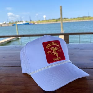 May include: White trucker hat with a red and gold Fireball Whisky logo patch. The logo features a red devil with a tail and the words "Red Hot Whisky".