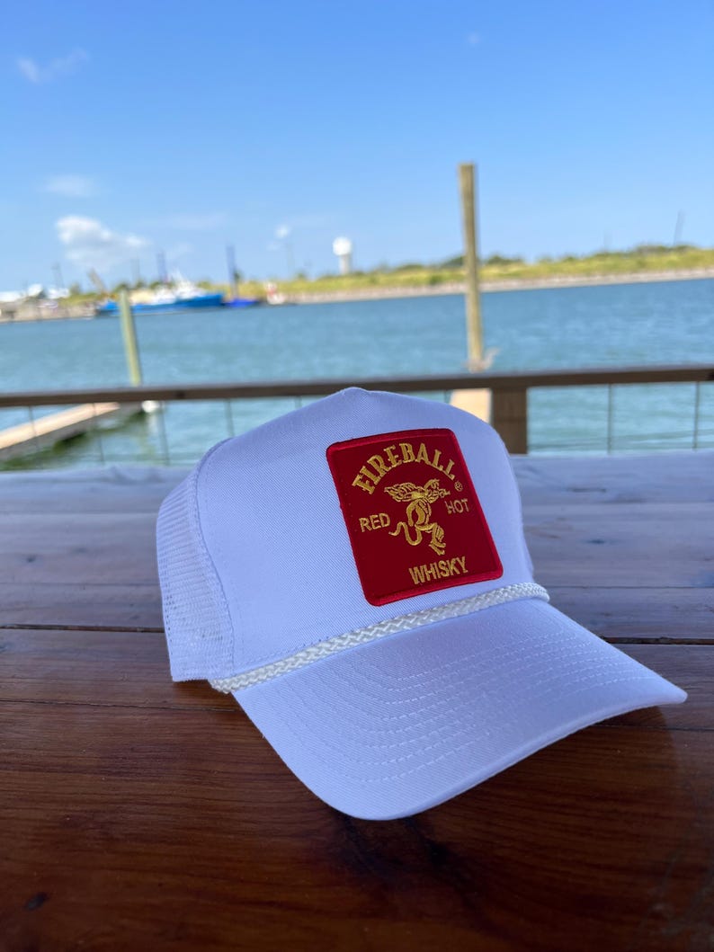May include: White trucker hat with a red and gold Fireball Whisky logo patch. The logo features a red devil with a tail and the words "Red Hot Whisky".