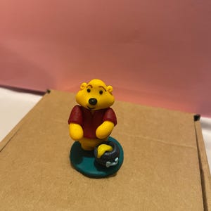 May include: A yellow bear figurine wearing a red shirt, standing on a teal base. The bear is next to a small black pot with yellow honey. The figurine is on a brown cardboard surface.