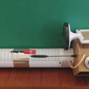 Könnte beinhalten: Ein einzigartiges, wissenschaftliches Gerät im Vintage-Stil mit einem weißen, zylindrischen Körper, der mit kleinen Quadraten bedeckt ist. Es verfügt über einen Holzkasten mit einem Bildschirm, der eine laufende Figur zeigt, ein Zifferblatt und einen weißen Rahmen. Rote und weiße Drähte gehen vom Gerät ab.