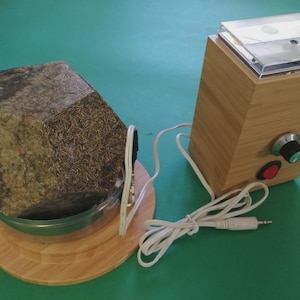 Puede incluir: Un dispositivo de madera con una parte superior transparente y un dial, conectado por un cable blanco a un objeto similar a una roca en un cuenco. El objeto similar a una roca está sobre una base de madera. El dispositivo tiene un botón rojo.